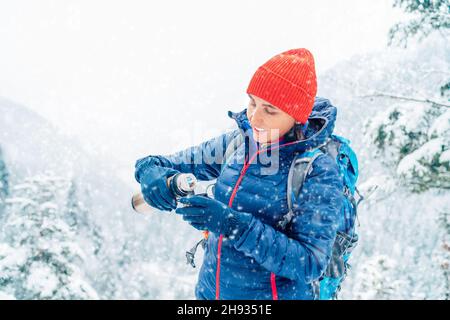 Smiling Woman dressed warm down jacket pouring a hot drink from thermos flask to mug while she has a break on trekking winter mountains route. Active Stock Photo