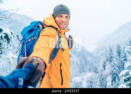Happy smiling Man dressed bright orange softshell jacket with backpack hand in hand girlfriend point view on the winter mountains route. Active people Stock Photo