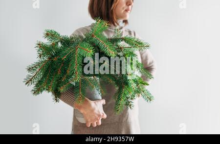 A woman dressed warm cozy sweater holding the clay vase full of green freshly cut fir-tree coniferous branches. Isolated on the white wall background Stock Photo