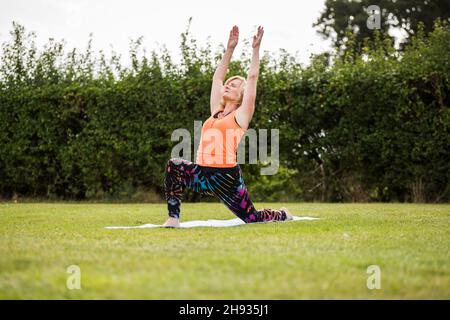 A middle aged woman practicing yoga barefoot outside in a grassy park. She  is wearing a bright orange vest and black leggings. The style of yoga she i  Stock Photo - Alamy