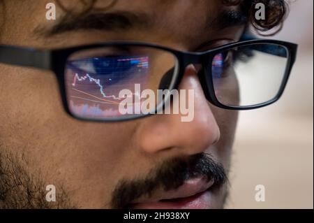 Close-up of Indian man, crypto trader wearing glasses, looking at computer screen with stock exchange market financial data charts reflecting in glasses Stock Photo