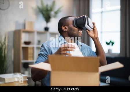 Happy african american man checking new VR headset while opening parcel box. Young male blogger doing reviews about new products for his subscribers. First person view. Stock Photo
