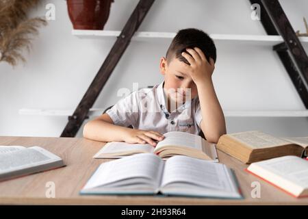 unmotivated student with headache sitting at home at his desk doing homework preparing for the lesson. Tired little school student reading books Stock Photo
