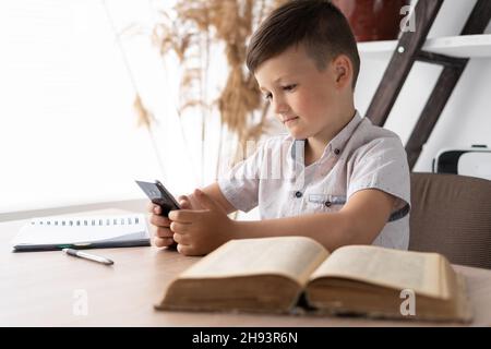 concentrated boy playing a game on the mobile phone while sitting at the table does homework. Gadget addicted student. Concept for online games or Stock Photo