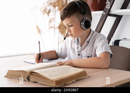 pupil Caucasian in headphones at the desk doing homework in a notebook reads a textbook. The child has a self-isolation lesson at home. Online Stock Photo