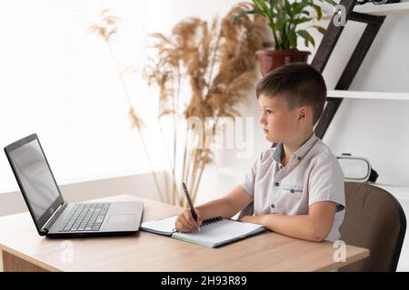 schoolboy boy studying at home doing homework with a laptop writing down the task in a notebook or notepad. junior elementary school student studies Stock Photo