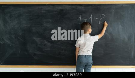 Back view of a child writes with chalk on a blackboard solution of an example in mathematics. Elementary school math lesson. Schoolboy boy thinks near Stock Photo