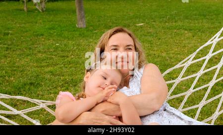 cute couple grandmother and granddaughter 3 years old are relaxing and basking in a hammock on a green meadow in their garden on a summer sunny day Stock Photo