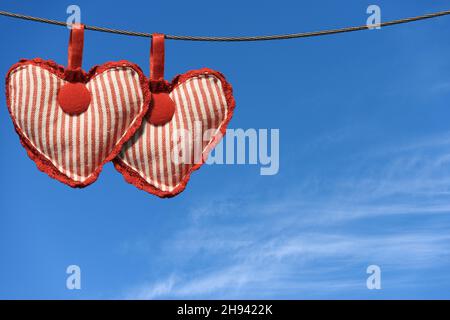 Close-up of two hearts made of cloth hanging on a steel cable against a blue sky with clouds and copy space. Stock Photo