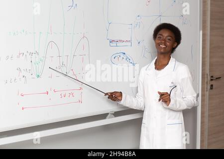 African American smiling woman math teacher stands at black board with pointer. Stock Photo