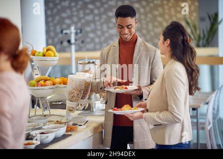 A young man is having a friendly conversation with female colleague while helping themselves with a food at the lunch break in a relaxed atmosphere du Stock Photo