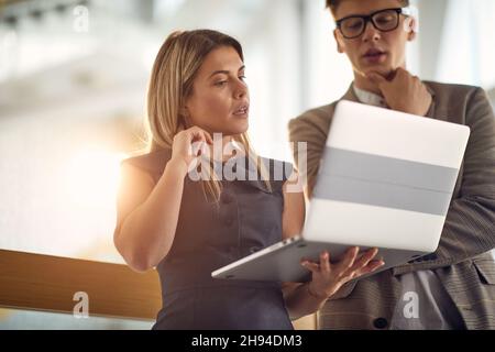 A young businesswoman and her young male colleague are standing in the hallway and they are focused on a laptop content they are watching together in Stock Photo