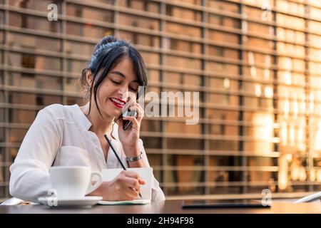 smiling businesswoman working and talking on the phone on the terrace of a coffee shop, concept of digital entrepreneur and urban lifestyle, copy spac Stock Photo