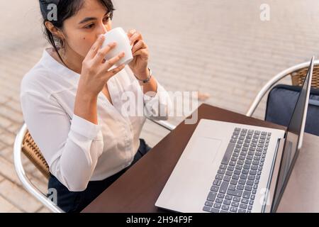 businesswoman drinking a cup of coffee while working on her laptop on a terrace, concept of digital entrepreneur and urban lifestyle, copy space for t Stock Photo