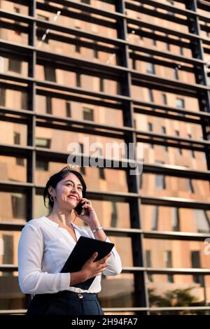 smiling businesswoman talking on the phone and using her tablet in front of an office building, concept of successful entrepreneur and urban lifestyle Stock Photo