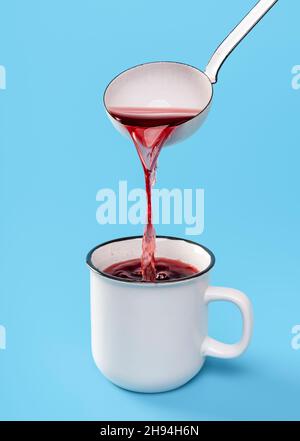 Pouring hot wine from a white ladle in an enamel mug on a blue table. Mulled red wine in a white cup, isolated on a colored background. Stock Photo