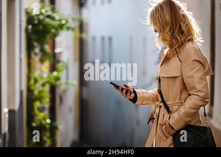 Woman with trench coat using her smartphone on city street. Woman text messaging on mobile phone Stock Photo