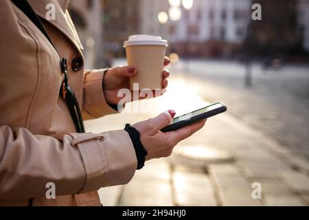 Businesswoman using mobile phone during city break on street. Woman text messaging on smartphone and drinking coffee Stock Photo