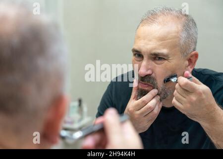 Serious handsome man looking in mirror and shaving his whiskers Stock Photo