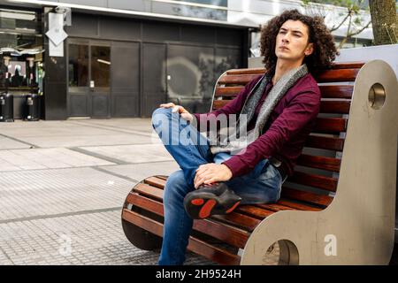 A Young curly haired man sitting on a bench in the street and waiting with serious expression. Stock Photo