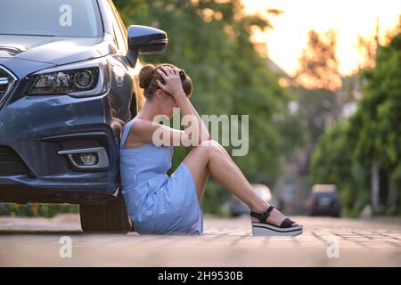 Young woman driver sitting beside her broken car waiting for help. Vehicle problems concept. Stock Photo