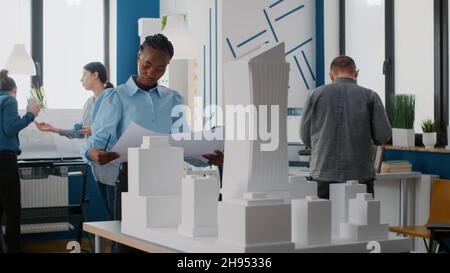 Portrait of african american architect looking at blueprints plans to work on building model. Woman engineer analyzing sketch to design maquette and constrution layout for project. Stock Photo
