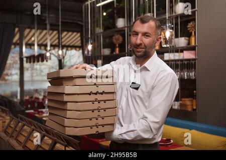 Male waiter holding many pizza boxes, walking through the restaurant Stock Photo