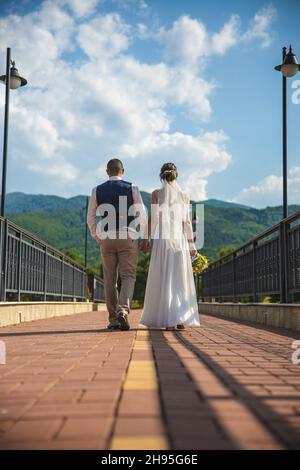 Circa 2021, Bulgaria. Newlyweds hold hands and run along the road. Wedding Day concept.Beautiful mountain and sky view. High quality photo Stock Photo