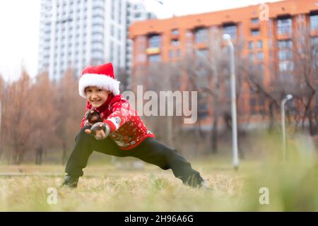 a boy in a warm Christmas sweater with a New Year's deer and a Santa hat performs stretching and gymnastic exercises while making faces Stock Photo