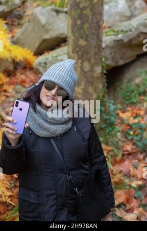 portrait of a woman with sunglasses and wool hat taking self-portraits with smartphone in the forest on an autumn day in the morning. Stock Photo