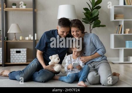Happy parents hugging cute daughter girl, watching child using smartphone