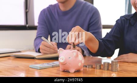saving concept the man in blue sweater writing income account while the female in dark blue shirt drop a coin into a piggy bank. Stock Photo