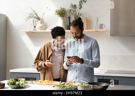 Happy young African American couple using cellphones, cooking in kitchen. Stock Photo