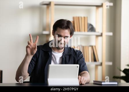 Positive millennial employee guy, tablet user waving hello at screen Stock Photo