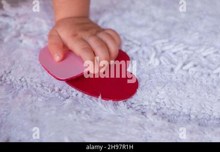 Baby's hand holds hearts on a white background. A child tries to grab hearts from a white fluffy carpet