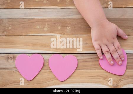 Children's hands move the heart to themselves on a wooden background. Three hearts on the surface. The child's hands are choosing. Stock Photo