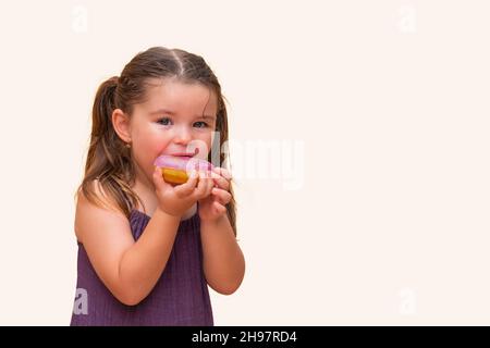 cute little girl in a dress bites a delicious, sweet, pink donut.  child smiles in anticipation of a harmful, tasty meal on a yellow orange background Stock Photo