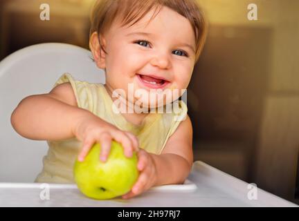 baby with an apple. baby with green apple on baby feeding chair in kitchen Stock Photo