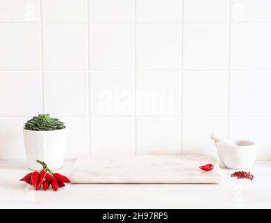 White kitchen countertop with white tiles, marble board for product presentation or advertising. Spices and cooking utensils on both sides. Front view Stock Photo