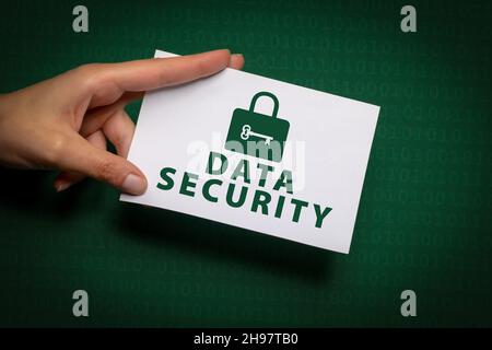 female hand holds a piece of paper with the inscription Data Security and lock symbol in green writing over dark green background with binary code Stock Photo