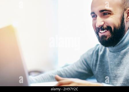 Young man with beard looks at laptop screen and smiles. Internet dating or communication through video communication. Young positive man of Arab appearance. Emotional Close-up Portrait. Stock Photo
