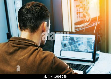 An IT specialist has been working on a laptop in a data center for 30-40 years, sitting near server equipment. Starting diagnostics or performing maintenance work. An authentic scene. View from the back Stock Photo