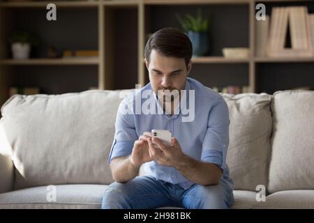 Focused millennial guy un casual using cellphone on couch Stock Photo