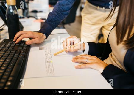 Close up of man's hands using his colleague's computer and woman's hand holding a pen, on a messy desk, in office. Working toghether Stock Photo
