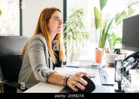 Young red haired woman, sitting on a desk and using a computer, in modern office Stock Photo