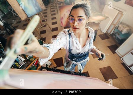 Artist painting while wearing tinted eyeglasses. Imaginative young painter making a new artwork for her project. Creative female artist standing in fr Stock Photo
