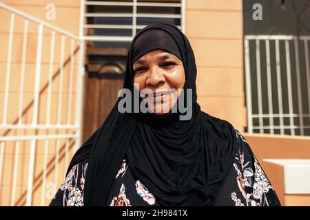 Cheerful Muslim woman smiling at the camera while standing in front of her home during the day. Self-confident middle aged woman wearing a hijab and s Stock Photo