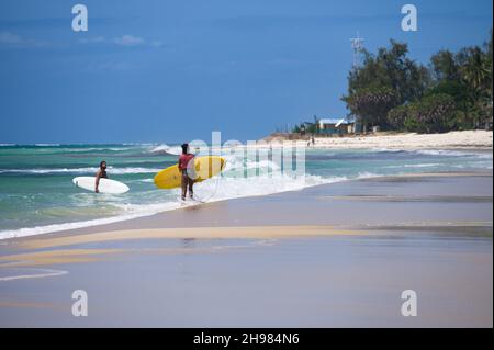 Two surfers walking out of the ocean as waves break on the beach, Diani, Kenya Stock Photo