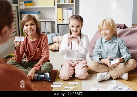 Group of happy children sitting on the floor and playing in English game together with teacher in the classroom Stock Photo