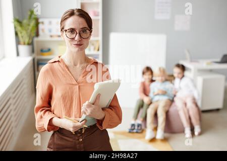 Portrait of young teacher in eyeglasses holding her report looking at camera while standing in the classroom with children in the background Stock Photo